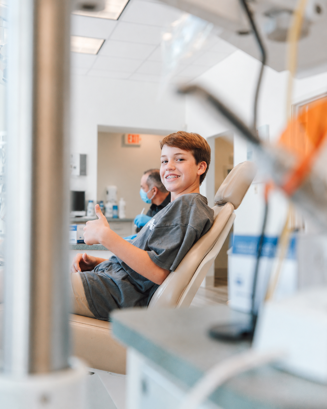 Boy with braces smiling and giving a thumbs-up in a dental chair; background staff edited out